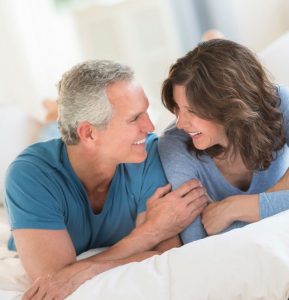 Model image showing a middle-aged man and woman lying in bed on stomach smiling at each other, depicting no sleep apnea, Charlotte, NC.